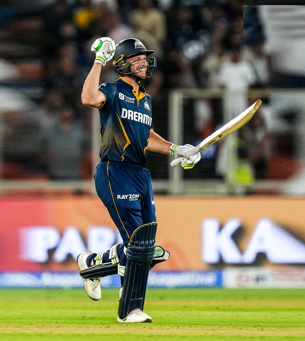 A cricket player in a blue jersey celebrates triumphantly while raising his bat, with a cheering crowd in the background.